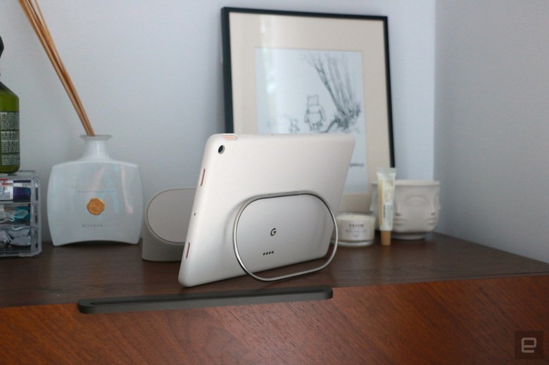 The Pixel Tablet on a walnut dresser propped up by the silver kickstand on the back of its protective case.
