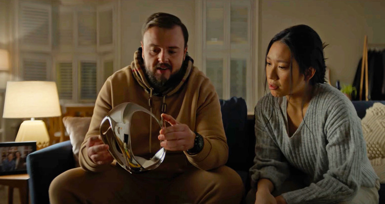 A man and woman sit on a living room couch and both look at a shiny object that the man is holding.
