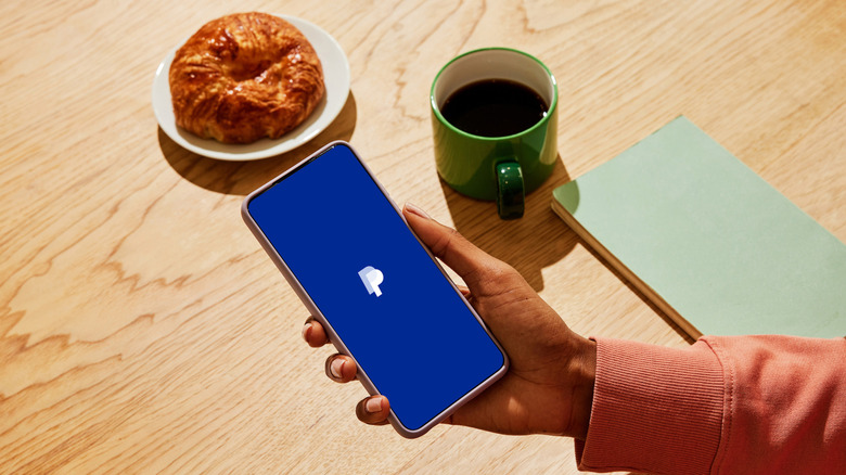 Photo looking down at a person's hand holding a smartphone with the PayPal app logo visible. Backdrop is a pale wooden table with a croissant, a cup of coffee and a slim notebook.