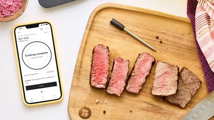 Top-down photo of a KitchenAid Smart Thermometer next to slices of cooked meats at different donenesses on a wooden cutting board. A smartphone to the left reads
